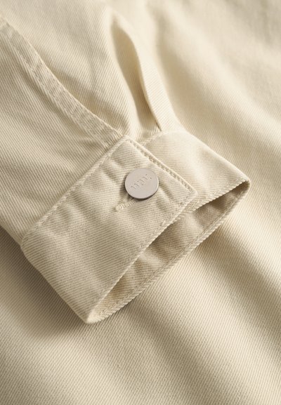 Artimas vaizdas į smėlio spalvos audinio rankovę su metaliniu spaudžiamu mygtuku, ant sulenkto rankogalių išgraviruotu „NN07“.