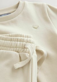 Ensemble en sweat-shirt et sweat-pantalon beige. Le sweat-shirt possède un col côtelé et un logo brodé. Le pantalon est doté d'une taille élastique avec cordon de serrage.