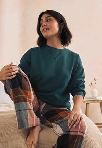 teal sweatshirt med rund hals; ternede, flerfarvede flannelbukser; siddende på et tekstureret, beige sengetæppe, lys baggrund.