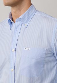 Camicia di colore azzurro chiaro con sottili righe verticali, colletto button-down e taschino sul petto con etichetta. Tessuto morbido e liscio.