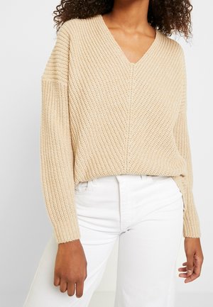 Beige gestrickter Pullover mit V-Ausschnitt, langen Ärmeln und strukturiertem Finish, kombiniert mit weißen Hosen. Auffällige Rippdetails an den Ärmeln.