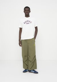 Λευκό μπλουζάκι με κόκκινη και μπλε εκτύπωση "ADIDAS 1994", συνδυασμένο με φαρδιά χακί παντελόνια και μπλε σκούρα αθλητικά παπούτσια με τρεις ανοιχτό μπλε ρίγες.