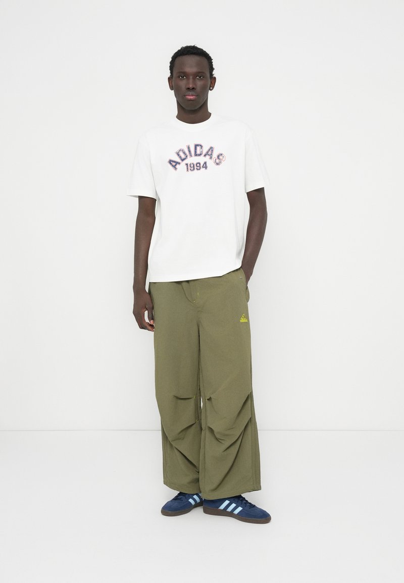 T-shirt blanc avec l'inscription rouge et bleue "ADIDAS 1994", porté avec un pantalon ample vert olive et des baskets bleu marine avec trois bandes bleu clair.