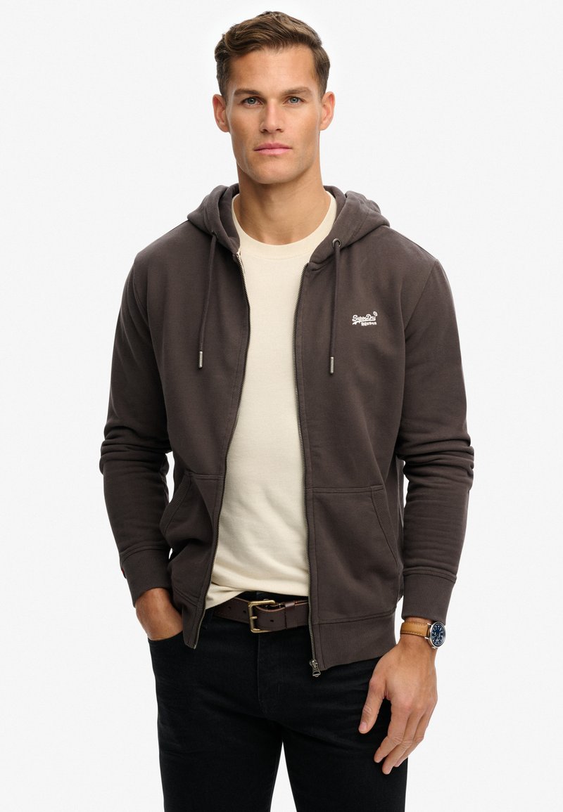 Braune Zip-Hoodie mit Kapuze und Zugband, ausgestattet mit einem weißen Logo auf der Brust und frontalen Taschen, kombiniert mit einem cremefarbenen T-Shirt und dunklen Jeans.