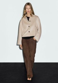 Beige wool-blend jacket with a collared neckline and four black buttons, paired with brown suede pants and black flat sandals.