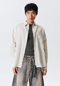 Young man wearing an open beige button-up shirt over a dark gray top and faded wide-leg jeans with a keychain hanging from the belt loop.