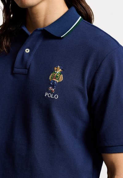 Granatowa koszulka polo z teksturowanej tkaniny; posiada zieloną i białą prążkowaną kołnierzyk oraz wyszywanego niedźwiedzia w kurtce i dżinsach.