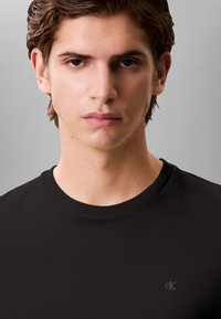 T-shirt preta de algodão com gola redonda e mangas curtas, com um pequeno logótipo cinzento no lado esquerdo do peito. Textura lisa.