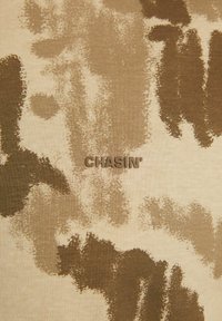 Tan ir rudos spalvos kamufliažo rašto audinys su iškilniu prekės ženklu "CHASIN'" centre. Lygus tekstūra, kasdienis dizainas.