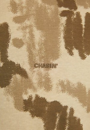 Tkanina w kolorze brązowym i beżowym w wzór kamuflażowy z wytłoczonym na środku logotypem "CHASIN'". Gładka tekstura, casualowy design.