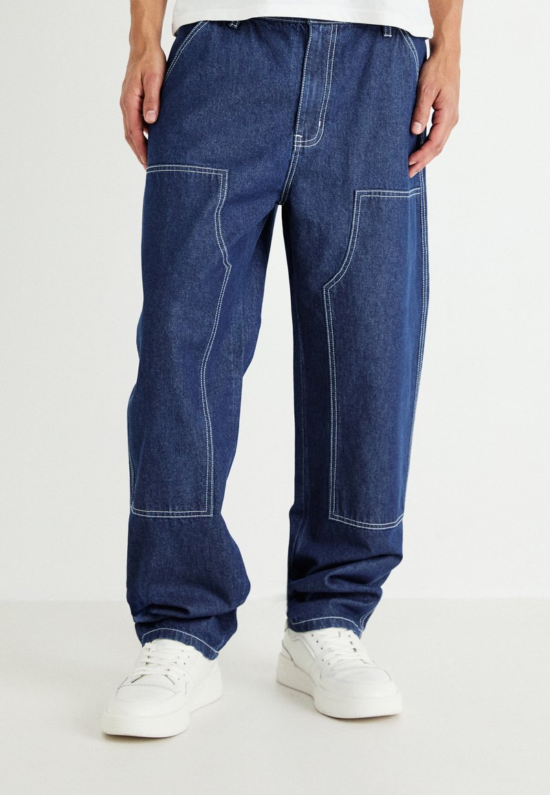 Sproščeno krojene jeans hlače v temno modri barvi s kontrastnim belim šivom, z obližno žepno in elastičnim pasom, v kombinaciji z belimi supergami.