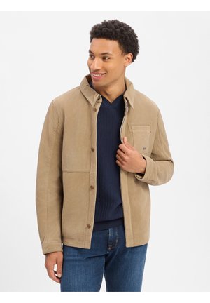 Mann lächelt, trägt eine hellbraune Jacke mit Knöpfen über einem dunkelblauen Pullover und blauen Jeans, vor schlichtem weißem Hintergrund.