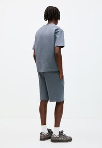 Set di t-shirt e pantaloni corti in cotone grigio con una vestibilità rilassata. I pantaloni corti presentano tasche laterali. Il completo è indossato con sneaker grigie e calzini bianchi.
