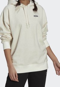 Osoba oblečená do světlé béžové mikiny Adidas s logem na přední straně, s kapucí na šňůrku a v černých kalhotách, zachycena od ramen po boky.