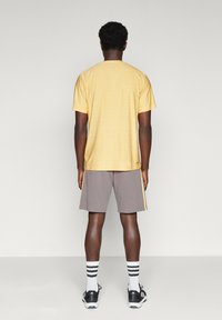 Gelbes Kurzarmhemd und graue Shorts mit gelben Seitenstreifen, kombiniert mit schwarzen Sportschuhen und gestreiften Socken.