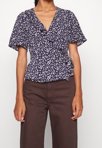 Blusa avvolgente floreale blu navy con bordi arricciati e maniche corte a sbuffo, realizzata in un tessuto leggero. Abbinata a pantaloni palazzo marroni.