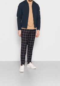 Námořnická modrá bomber bunda přes béžovou košili, doplněná černými kostkovanými kalhotami. Oblečení doplňují bílé tenisky.