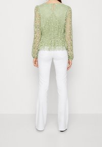 Top in pizzo verde chiaro con maniche lunghe, punto vita peplum e dettaglio a chiave sul retro, abbinato a pantaloni bianchi a campana. Tessuto liscio e motivo floreale.
