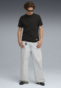 Camiseta de algodón negro con logo tonal, combinada con jeans de pierna ancha en azul claro. Llavero colgando del bolsillo, usada con zapatos negros sin cordones.
