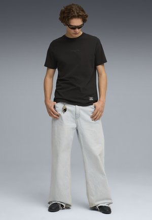 Camiseta de algodón negro con logo tonal, combinada con jeans de pierna ancha en azul claro. Llavero colgando del bolsillo, usada con zapatos negros sin cordones.
