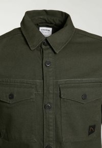 Chemise boutonnée vert foncé avec deux poches poitrine, boutons noirs et col, affichant un petit patch logo noir et orange sur une poche.