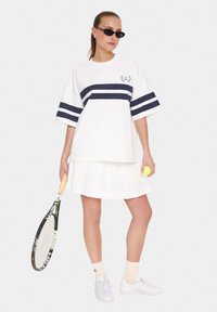 Camiseta blanca de gran tamaño con rayas marinas, combinada con una falda blanca con plisados. El modelo sostiene una raqueta de tenis y una pelota de tenis de color amarillo brillante.