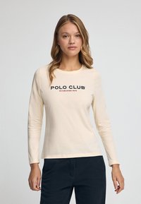 Cremefarbendes Langarmshirt mit rundem Ausschnitt. Verfügt über das "POLO CLUB"-Logo in dunkelblauem und rotem Text. Aus weichem, glattem Baumwollstoff. Minimalistisches Design.