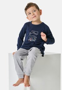 Marineblaue Langarmoberteil mit Berggrafik und "EXPLORE"-Text, kombiniert mit grauen Sweatpants mit Umschlag. Das Kind sitzt auf einem weißen Block.