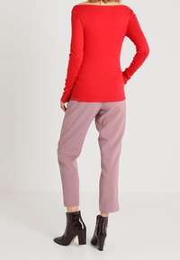 Rotes Langarmoberteil mit Off-Shoulder-Design, kombiniert mit helllila Hosen. Das Model trägt glänzende schwarze Stiefeletten mit einem klobigen Absatz.