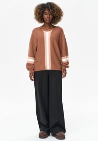 Vrouw met krullend haar die een bruine trui met verticale strepen, zwarte wijd uitlopende broek en bruine schoenen draagt, staand tegen een witte achtergrond.