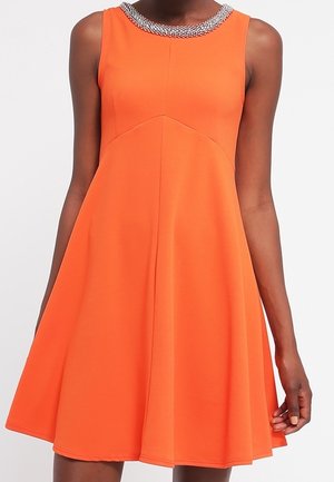 Mouwloze oranje A-lijn jurk met een met kralen versierde halslijn, gedragen door een persoon die staat tegen een effen achtergrond.