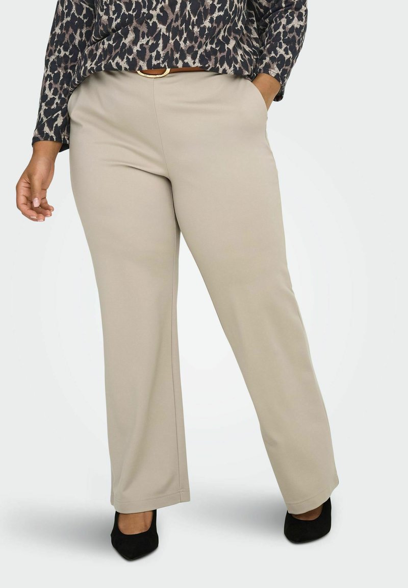 Bež hlače širokih nogavica glatke teksture, s mid-stručnim strukom i džepovima, uparene s crnim cipelama šiljastog vrha.