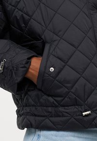 Prešívaná čierna puffer bunda s patentkami a elastickými manžetami. Tkanina má hladký povrch s mierne lesklým efektom.