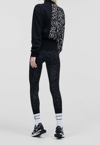 Sweatshirt noir avec un dos à motifs, associé à des leggings noirs avec un design géométrique. Porté avec des chaussures de sport noir et blanc.