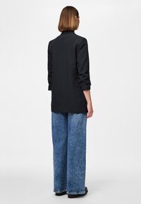 Blazer preto com gola alta, ajuste sob medida e mangas longas, combinado com jeans de ganga azul claro de perna larga e sapatos pretos.