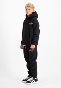 Zwarte hoodie met voorzak en logo, gecombineerd met bijpassende joggers en zwarte sneakers met witte strepen. Glad materiaal, casual fit.