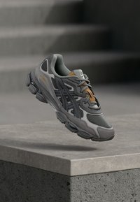 Γκρι αθλητικό παπούτσι με μείγμα διχτυωτού και συνθετικού υλικού. Διαθέτει υφή στην επιφάνεια και αντιθετικούς πορτοκαλί τόνους στη γλώσσα.