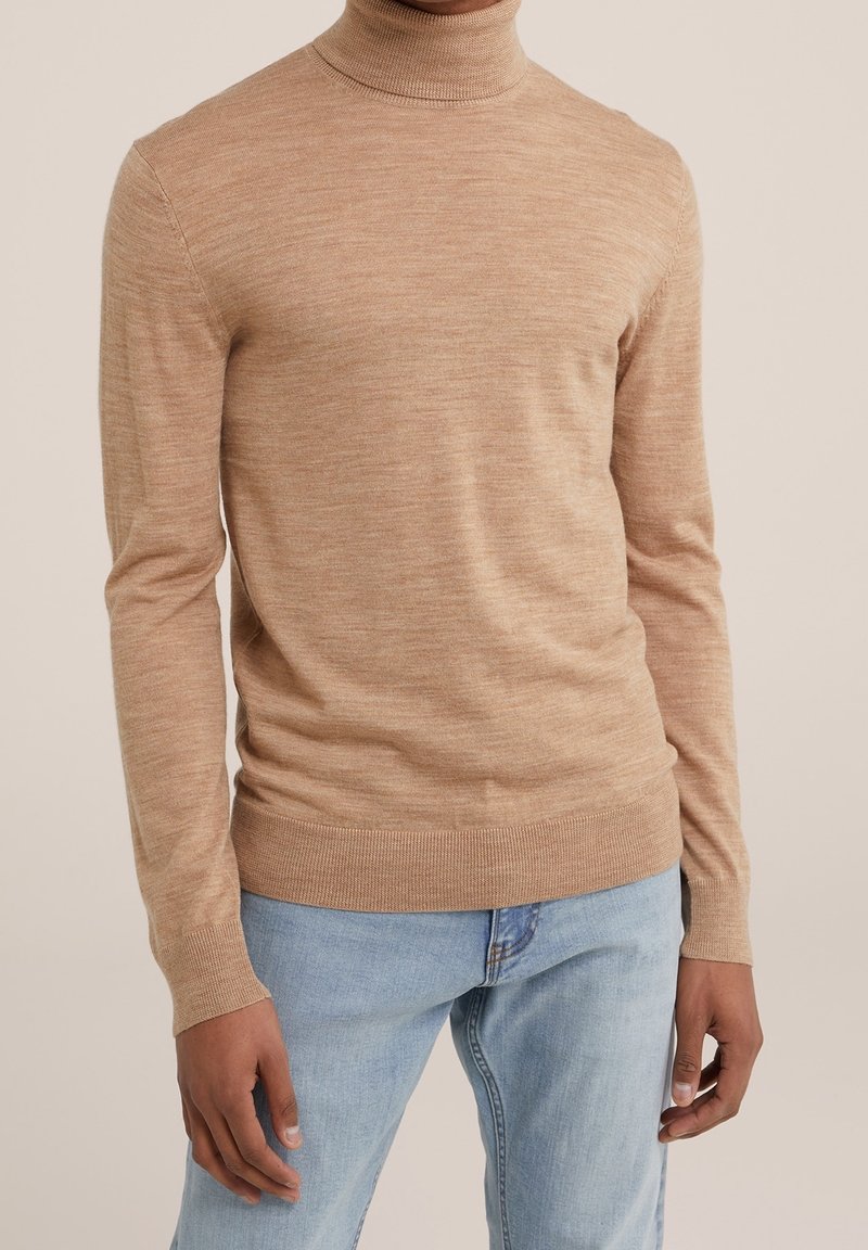 Un mannequin porte un pull à col roulé beige à manches longues, avec des poignets et un bas côtelés, associé à un jean bleu clair.