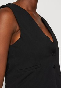 Kvinna klädd i en ärmlös svart kavaj med två knappar och en smal ficka på bröstet mot en neutral bakgrund.