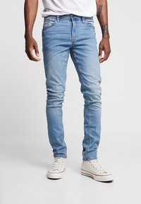 Světlé modré džíny slim fit z denimu s vybledlou texturou a jemným popraskaným efektem. Kombinovány s bílými teniskami pro kontrast.
