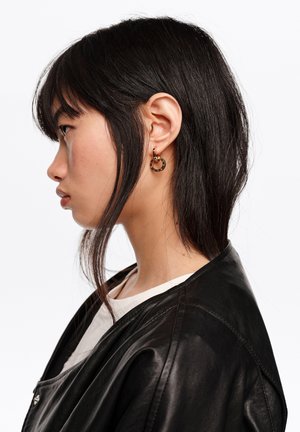 Młoda kobieta o czarnych włosach w profilu, nosząca złote kolczyki koła oraz czarną skórzaną kurtkę na białej bluzce, na jednolitym tle.