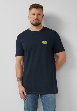 Mężczyzna z krótkimi włosami i brodą, ubrany w ciemnoniebieską koszulkę z żółtym logo Michigan oraz jasnoniebieskie dżinsy, stoi na tle gładkiej ściany.