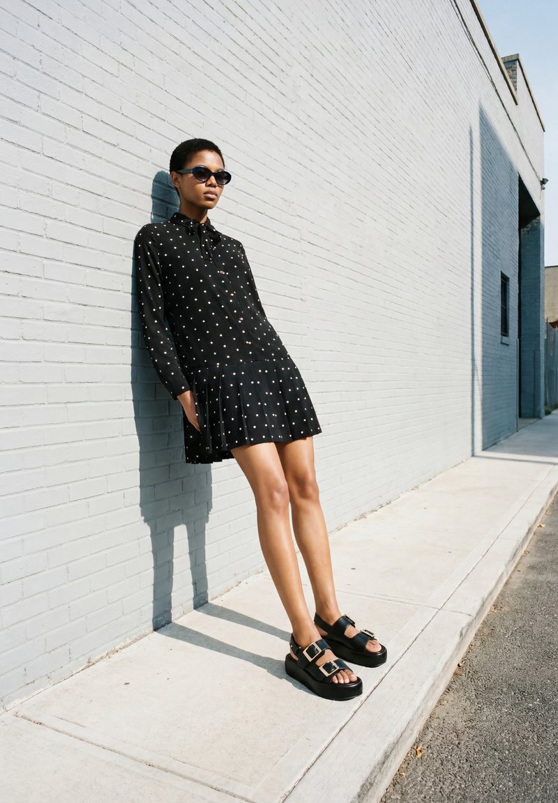 Personne portant des lunettes de soleil, une robe noire à pois et des sandales noires, adossée à un mur de briques gris clair sur un trottoir ensoleillé.