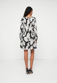 Melnā kleita ar baltu lapu rakstu, garām piedurknēm, pleatēta tekstūra, atslēgas cauruma aizmugures detaļa, un valkāta ar melniem augstpapēžu apaviem.