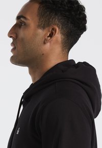 Zwarte hoodie met een zachte textuur, voorzien van een trekkoordcapuchon en een klein geborduurd logo op de borst. Zijaanzicht van een model.