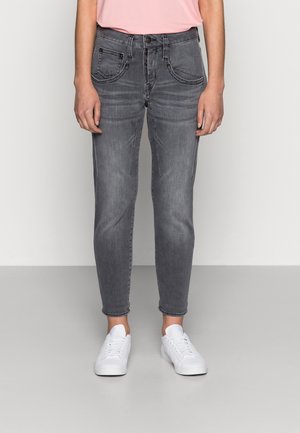 Osoba w wyblakłych szarych obcisłych dżinsach, różowej koszuli i białych trampkach, stojąca na jednolitym jasnym tle.