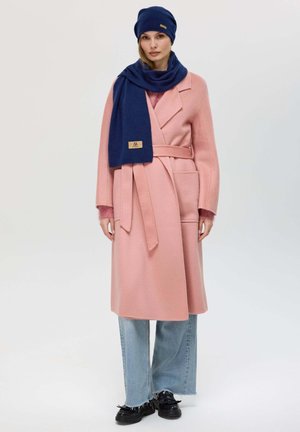 Cappotto in lana rosa chiaro con cintura a fiocco, due tasche frontali, abbinato a una sciarpa e un berretto blu navy; il modello indossa jeans azzurri strappati e scarpe nere.