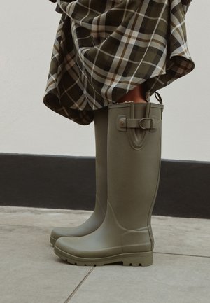 Bottes en caoutchouc vert olive montant jusqu'au genou, portées avec une jupe à carreaux marron et blanche, debout sur un sol carrelé près d'un mur blanc.