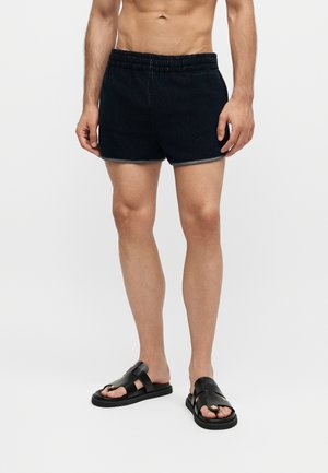 Homme portant un short en tissu éponge noir et des sandales en cuir noir, debout sur un fond blanc.