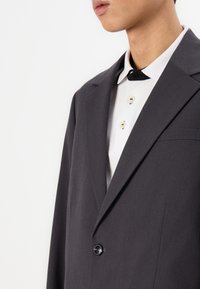 Donkergrijze blazer met een enkel knoopje, scherpe revers en een borstzak, gedragen over een witte blouse met kraag en zwarte accenten en knopen.